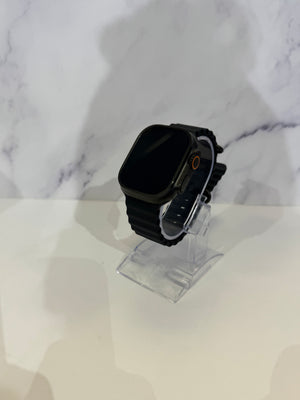A black smartwatch with a rectangular black screen and black strap, displayed on a clear acrylic stand against a white marble-patterned background.
