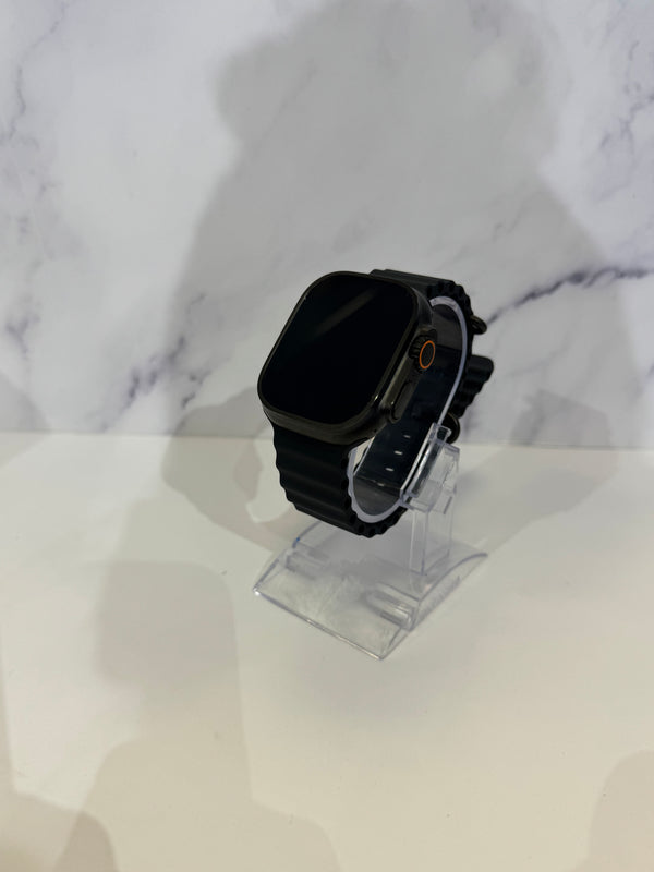 A black smartwatch with a rectangular black screen and black strap, displayed on a clear acrylic stand against a white marble-patterned background.