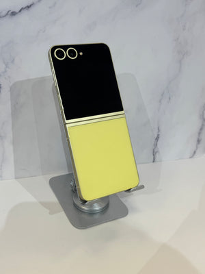 A foldable smartphone with a black top half and pale yellow bottom half, displayed upright on a clear stand against a white marble-patterned background.