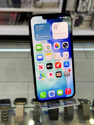 A black iPhone with its screen turned on, displaying the home screen with various app icons, photographed in a retail or showroom setting with glass display cases visible in the background.