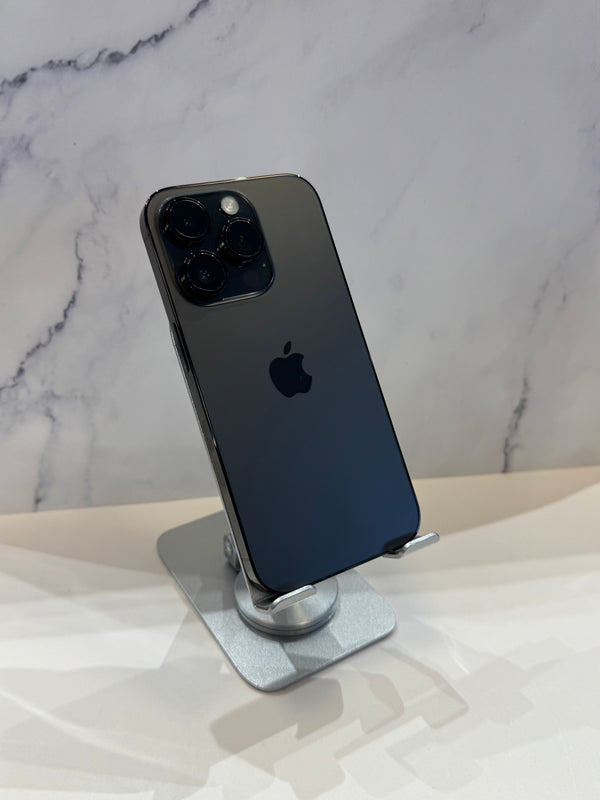 A black Apple iPhone 12 Pro Max standing upright on a silver metal display stand, showing the back of the phone with its triple-camera array and Apple logo.