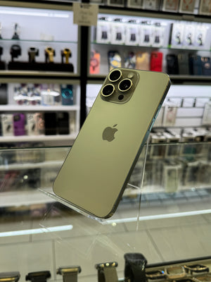 A gold-colored Apple iPhone shown from the back, resting on a glass display case inside a retail store. The Apple logo is visible in the center, and the background shows blurred store fixtures and other products.