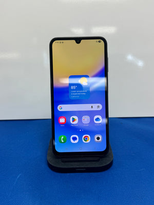 A black smartphone with a front-facing screen display resting on a black charging dock against a white wall and blue surface.