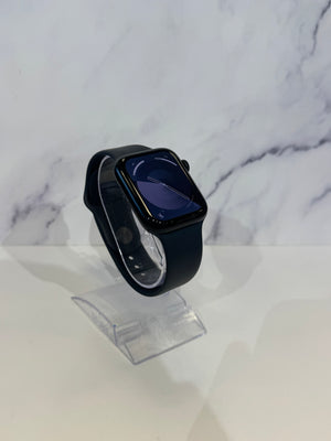 A black Apple Watch with a dark blue watch face and black sport band, displayed on a clear acrylic stand against a white marble-patterned background.