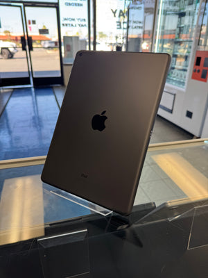A dark gray Apple iPad Pro standing upright on a clear display stand inside a retail store, with glass doors and vending machines visible in the background.