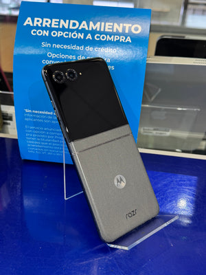 A Motorola flip-style smartphone shown open, with a black upper half and a gray lower half, displayed on a clear stand on a blue surface in front of a blue promotional sign.
