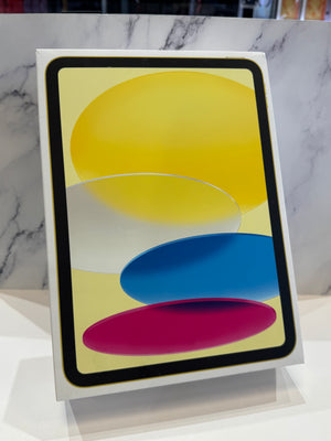 A tablet device in a white retail box, shown standing upright on a display stand against a white marble-patterned background. The screen displays an abstract design with overlapping yellow, blue, pink, and white shapes.