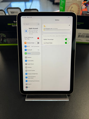 An iPad standing upright on a clear display stand, showing the Settings app screen with battery-related options. The device is photographed on a dark tabletop with blurred retail boxes in the background.