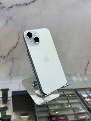 A light blue Apple iPhone 12 Pro shown from the back, resting on a silver metal display stand in a retail setting with a marble-patterned wall behind it.