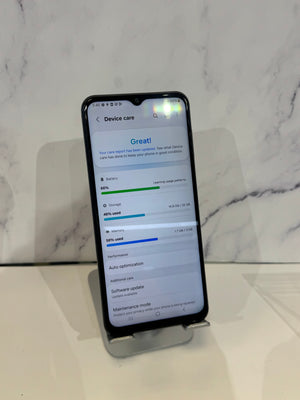 A black smartphone displayed upright on a small stand against a white marble-patterned background, showing a device care screen on the home screen.