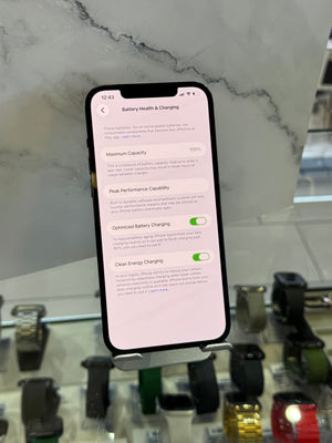 A black smartphone lying on a white marble surface, displaying the battery health and charging settings screen. The background shows a blurred retail display with various smartwatches or fitness trackers.