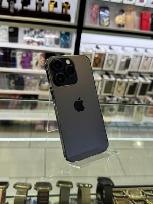 A dark gray Apple iPhone Pro shown from the back, standing on a clear acrylic display stand inside a retail store with shelves of other phones and accessories in the blurred background.