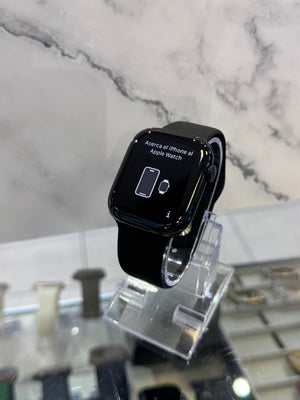 A black Apple Watch with a black band, displayed on a clear acrylic stand in a retail setting against a white marble-patterned wall.