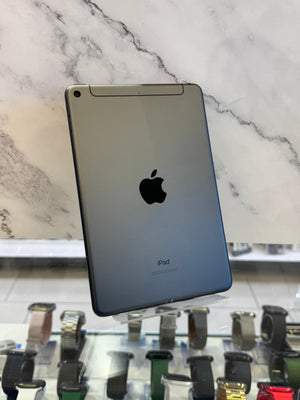 A silver Apple iPad shown from the back, standing on a clear display stand in front of a white marble-patterned wall. Several smartwatches are visible on a counter below.