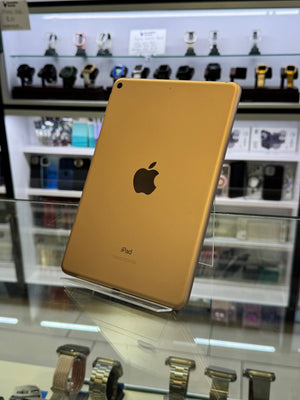 A gold Apple iPad shown from the back, resting on a glass display stand inside a retail store. The Apple logo and 'iPad' text are visible on the back of the device, with shelves of watches in the blurred background.