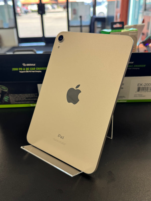 A gold Apple iPad shown from the back, standing upright on a clear display stand on a dark tabletop. The Apple logo is visible in the center, with retail packaging boxes blurred in the background.