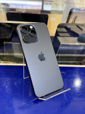 A gray Apple iPhone 12 Pro shown from the back, resting on a clear display stand against a blue reflective surface.