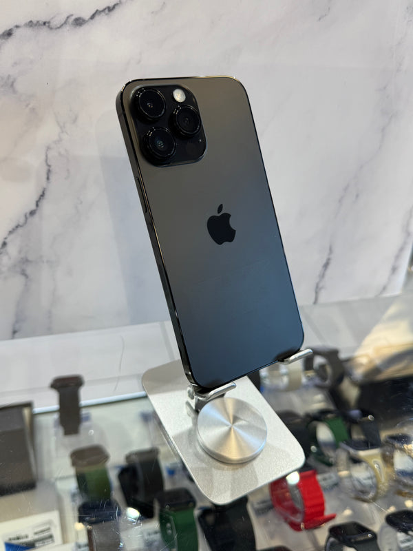 A dark gray Apple iPhone 12 Pro shown from the back, resting on a silver metal display stand in front of a white marble-patterned wall.
