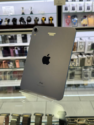 A silver Apple iPad shown from the back, standing on a clear display stand inside a retail store with shelves of accessories and watches blurred in the background.