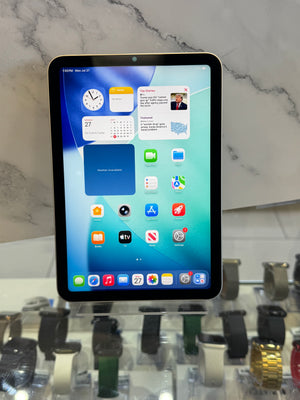 A tablet computer with a black bezel and silver edges, displaying its home screen on a white marble countertop, with a blurred watch display in the background.