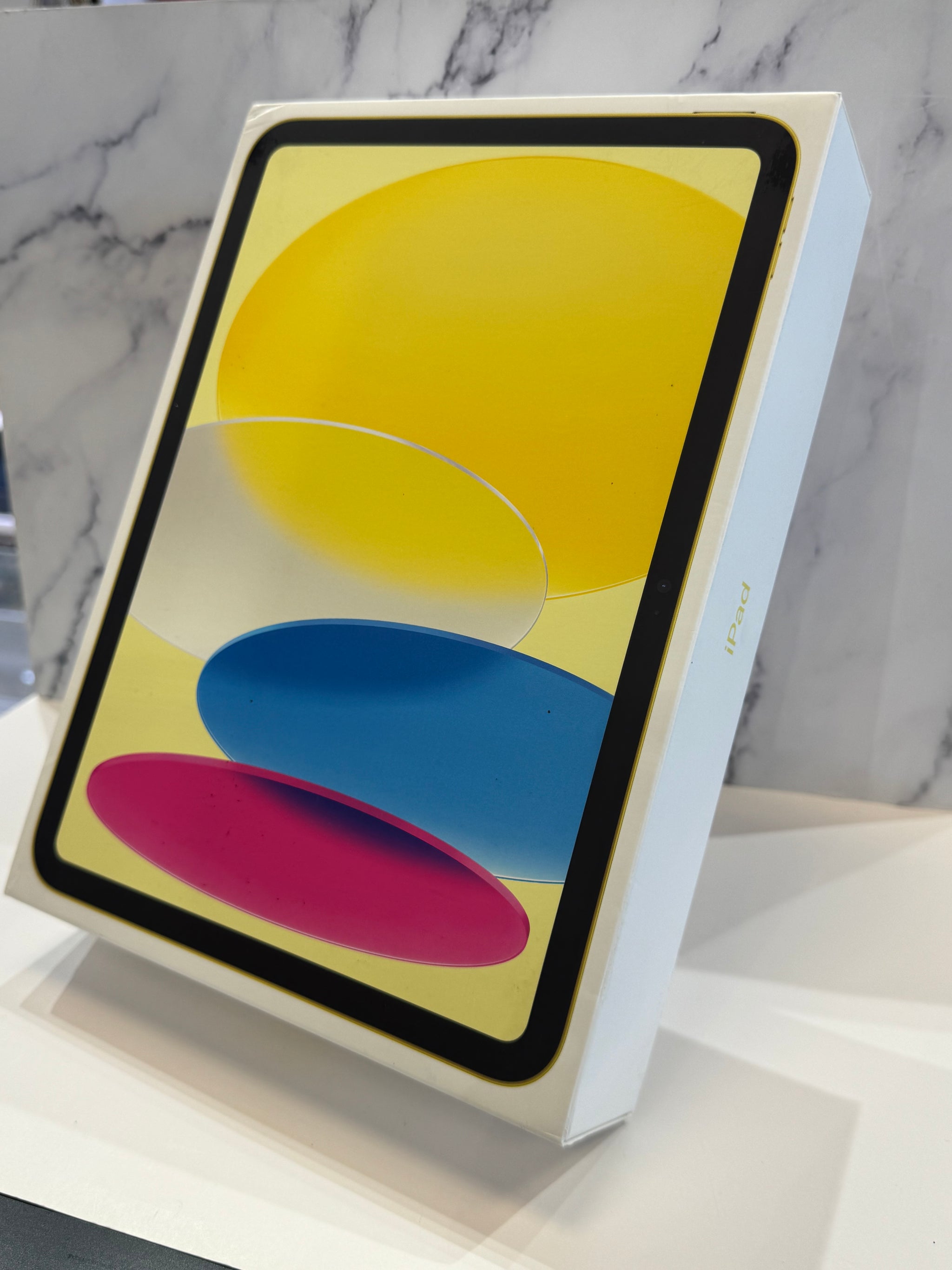 An Apple iPad box shown at an angle on a white marble-patterned surface, with the front display featuring a colorful abstract design in yellow, blue, and pink.