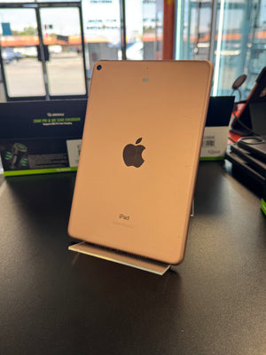 A gold Apple iPad standing upright on a small display stand on a dark tabletop, with a blurred store or office background and the Apple logo visible on the back.
