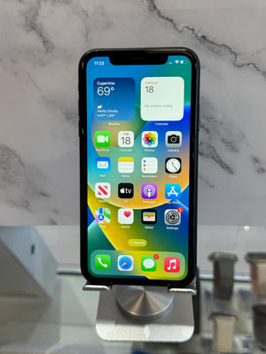 A black iPhone with its screen on, displaying the home screen, resting on a silver metal display stand against a white and gray marble-patterned background.