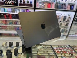 A closed Apple MacBook laptop in dark gray sits on a display stand inside a retail store, with watch and accessory displays visible in the background.