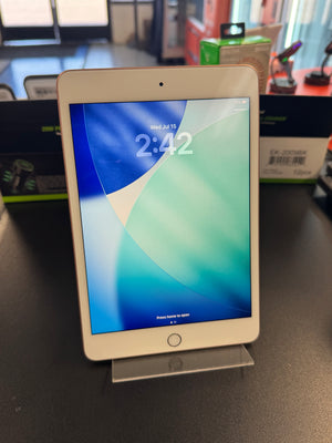 A white Apple iPad tablet standing upright on a small metal display stand on a dark tabletop, with its screen turned on and showing the lock screen.