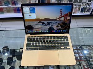 A gold Apple MacBook laptop with a black keyboard and trackpad, open and displaying its desktop screen, set on a glass display counter in a retail store.