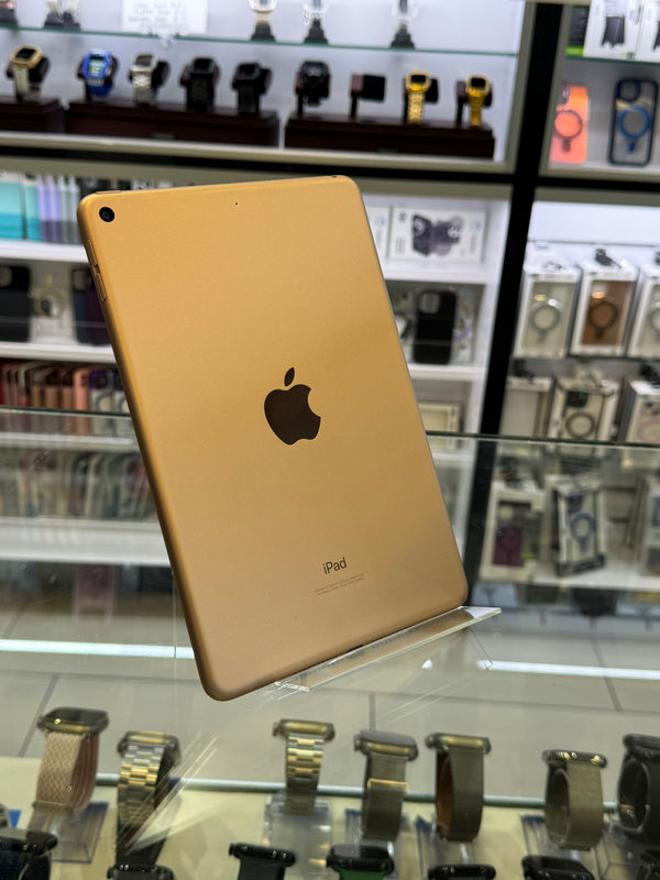 A gold-colored Apple iPad shown from the back, resting on a glass display counter inside a retail store with other electronics visible in the background.