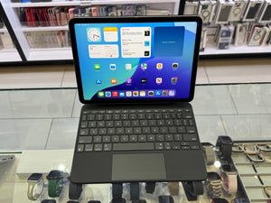 A black iPad with a matching keyboard case and trackpad, displayed on a glass counter in a retail store. The screen is turned on and displays various app icons and widgets.
