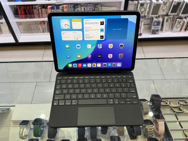 A black iPad with a matching keyboard case and trackpad, displayed on a glass counter in a retail store. The screen is turned on and displays various app icons and widgets.