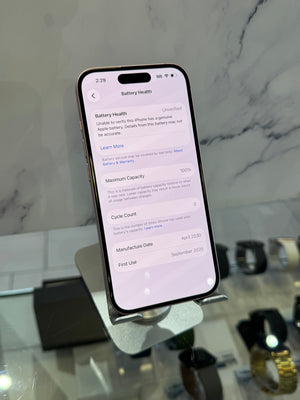 A black smartphone displayed upright on a stand in a retail setting, showing the Battery Health settings screen on its screen. The background features a gray marble-patterned wall and blurred watch displays on a counter.