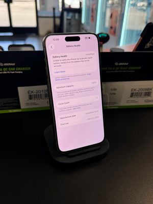A smartphone displaying battery health settings on a black charging stand, with a blurred retail or office background and green product boxes behind it.