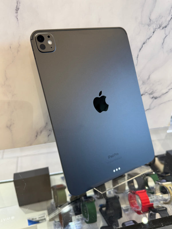 A dark gray Apple iPad Pro shown from the back, standing on a glass display case against a white marble-patterned wall.