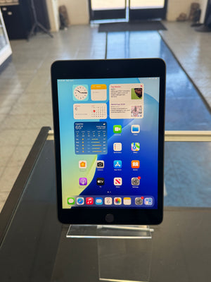 A black tablet device with its screen on, displaying the home screen with various app icons, photographed indoors against a gray tiled wall and counter.