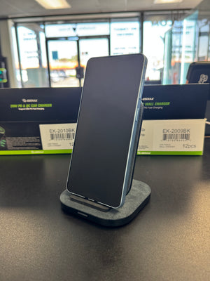 A black smartphone displayed upright on a gray charging dock, set on a dark tabletop with boxed charging products in the background.