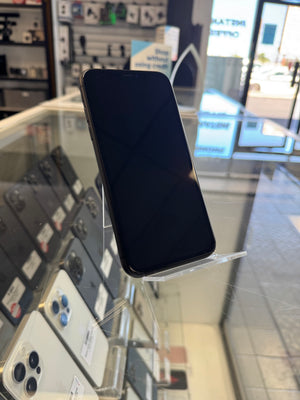 A black smartphone with a blank screen is displayed on a clear stand inside a retail store, with other phones and store fixtures visible in the background.