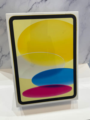 An Apple iPad in its original white box, showing a colorful abstract design on the screen with yellow, blue, and pink shapes, set against a marble-patterned background.