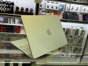 A closed Apple MacBook laptop is displayed on a glass counter inside a retail store, with shelves of accessories and watches blurred in the background.