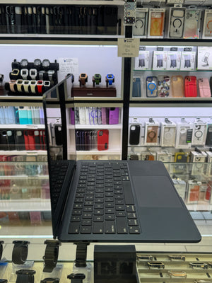 A black convertible laptop with its screen open and angled, displayed on a stand inside a retail store. The background shows shelves filled with various accessories, including phone cases, smartwatches, and earphones.