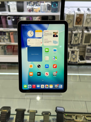 A tablet computer with a black bezel and colorful home screen interface, photographed inside a retail display area with shelves of watches or electronics in the background.