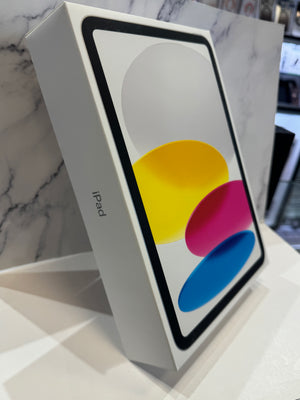 An Apple iPad box standing upright on a white surface, with the iPad screen graphic visible on the front and the word 'iPad' printed on the side. The background is a white marble-patterned wall.