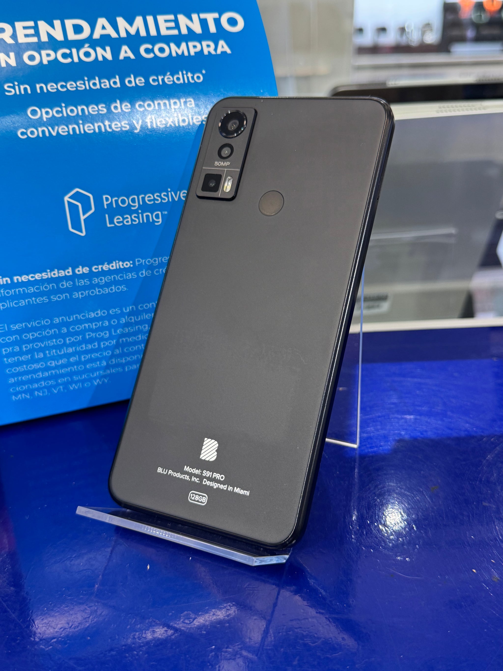A black Google Pixel Pro smartphone standing upright on a clear display stand on a blue table, with a blue leasing sign in the background.