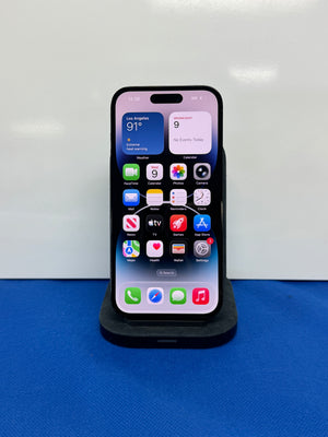 A black iPhone with its screen on, resting on a black charging dock against a white background with a blue surface below.
