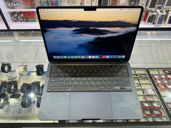 A gray Apple MacBook laptop with an open screen displaying a mountain mist wallpaper, set on a glass display counter with watches and accessories in the background.