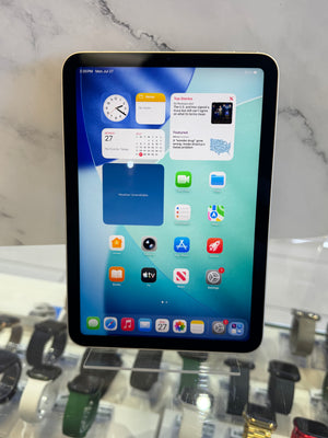 A tablet computer with a black bezel and a colorful home screen, resting on a white marble-patterned surface with blurred watch displays in the background.