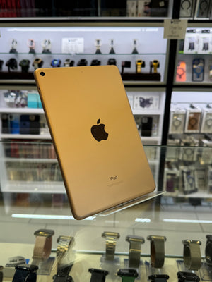 A gold Apple iPad shown from the back, resting on a glass display counter inside a retail store with shelves of other electronics in the background.