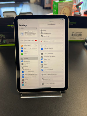 An Apple iPad displaying the Settings app on a clear stand on a dark tabletop, with a blurred retail environment in the background.
